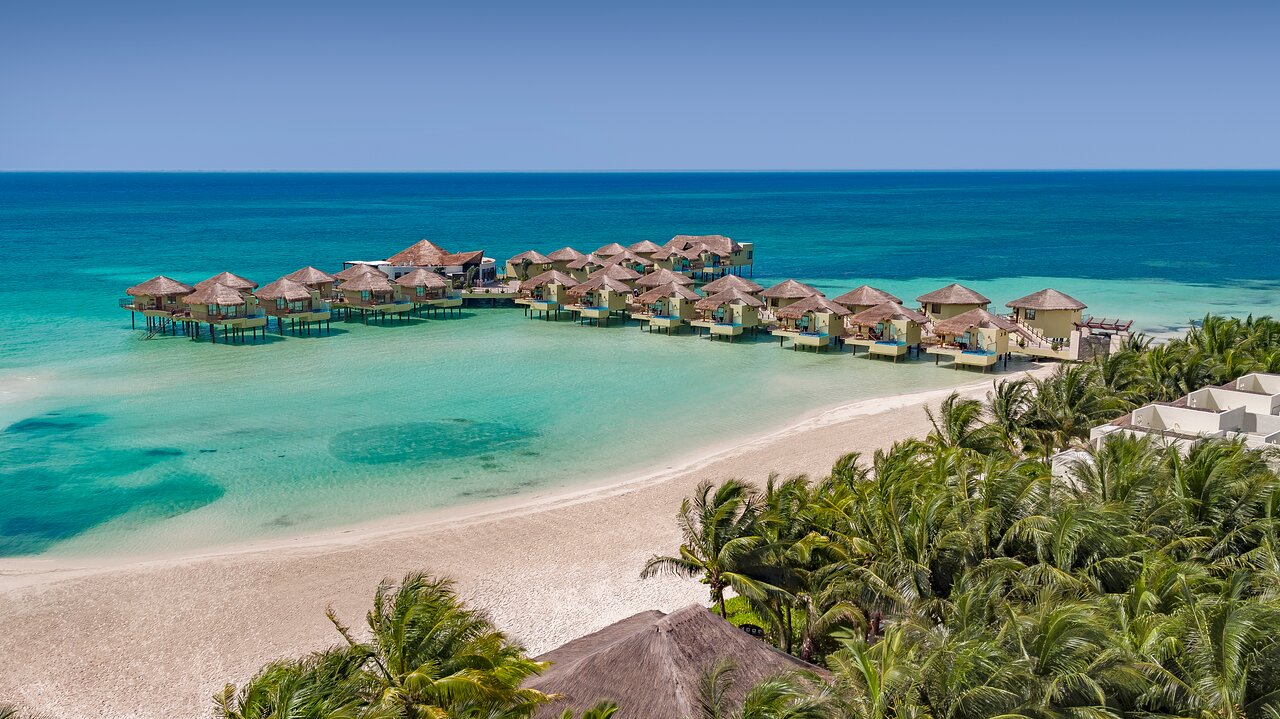 Vista aérea de Palafitos Overwater Bungalows en Riviera Maya, búngalos de lujo sobre el mar Caribe junto a playa y vegetación tropical, experiencia exclusiva para parejas.
